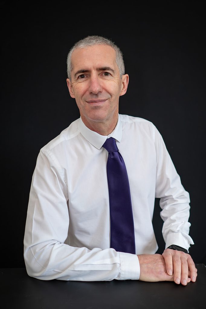 Business headshot in a white shirt and purple tie against a black background